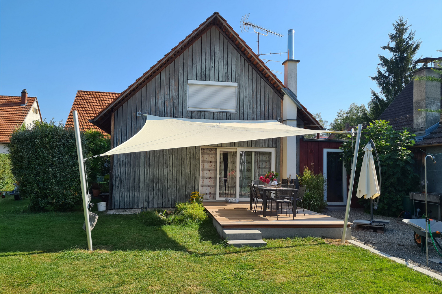 Cremefarbenes Sonnensegel vor kleinem Holzhaus und grüner Wiese